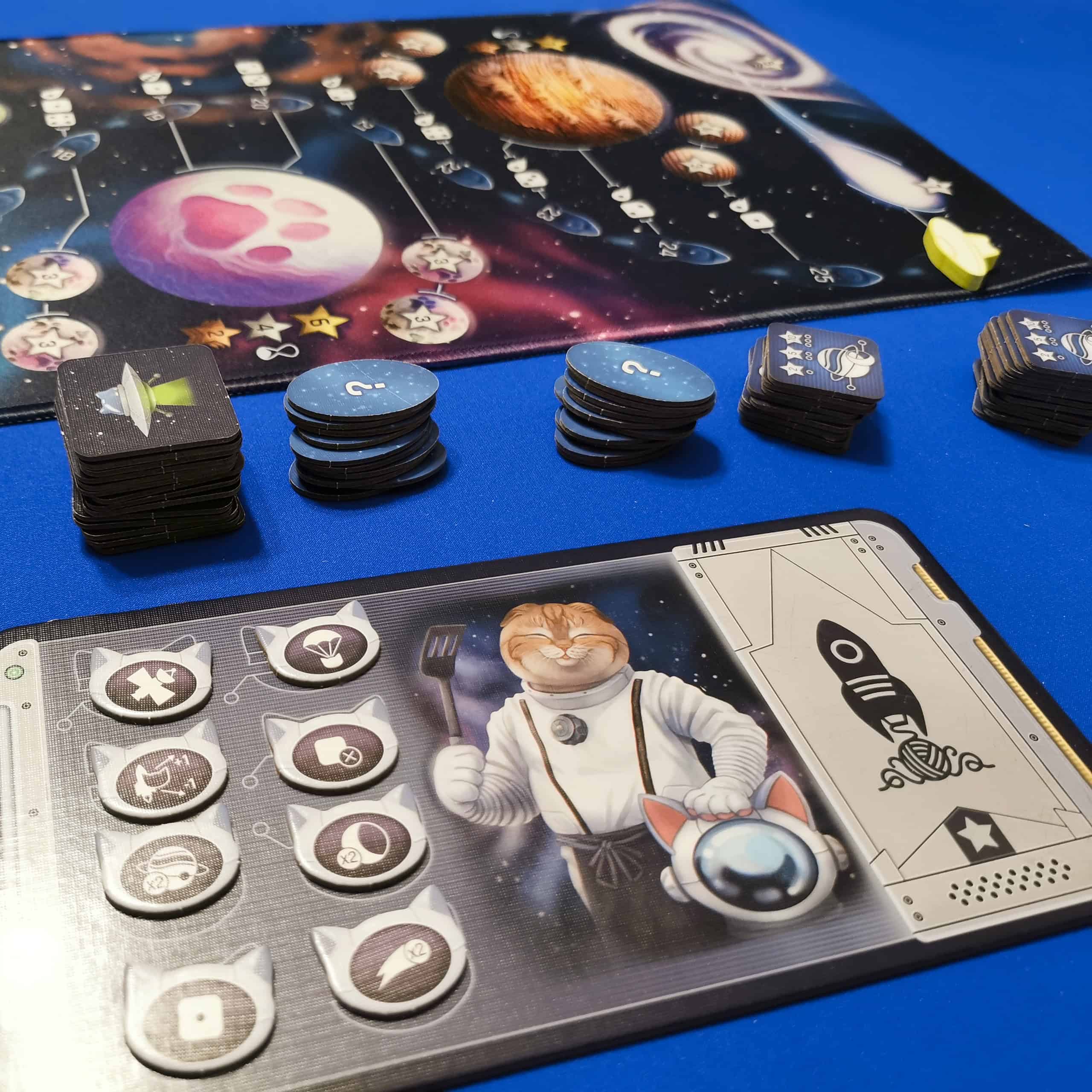 Kehittyneempien sääntöjen komponentteja, taustalla pelilauta ja edessä pelaajalauta, jossa kaikki kissa-astronautit.